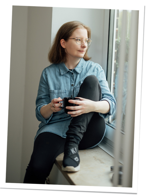 Eine Frau sitzt entspannt auf einer Fensterbank und hält eine schwarze Tasse in beiden Händen. Sie trägt ein Jeanshemd, eine Brille und schwarze Hosen. Ihr Blick geht nachdenklich aus dem Fenster.