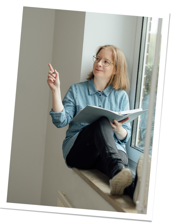 Eine Frau sitzt auf einer Fensterbank, trägt eine Brille und ein blaues Jeanshemd. Sie hält ein aufgeklapptes Notizbuch in der Hand und deutet mit der anderen Hand nach oben, als ob sie eine Idee hat.