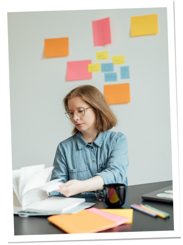 was-braucht-es Eine Frau in einem Jeanshemd blättert in einem Notizbuch auf einem Schreibtisch. Auf dem Tisch liegen bunte Haftnotizen, Stifte und eine schwarze Tasse. Im Hintergrund hängen Haftnotizen an der Wand.