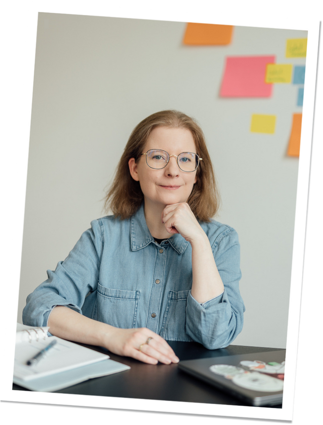 worauf kommt es an Eine Frau mit schulterlangem, hellbraunem Haar sitzt an einem Schreibtisch. Sie trägt eine Brille und ein Jeanshemd. Im Hintergrund sind bunte Haftnotizen an der Wand angebracht.
