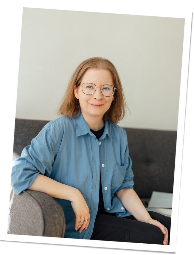 Eine Person mit Brille und blauer Bluse sitzt auf einem Sofa und schaut freundlich in die Kamera.